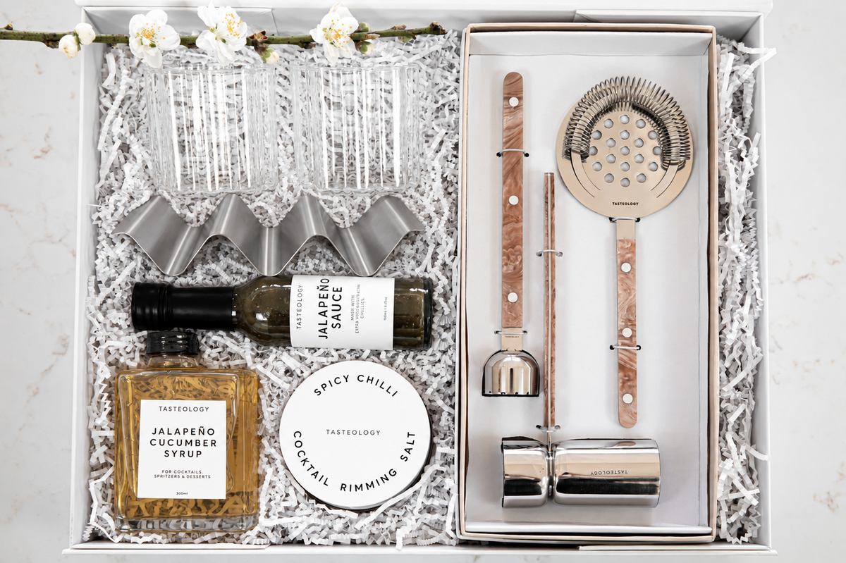 A curated gift box set from Tasteeology on a light marble surface. The open white box is filled with white paper shred and contains a bottle of jalapeño cucumber syrup, a round tin of spicy chili cocktail rimming salt, a set of clear textured glasses, a silver cocktail strainer, a wooden-and-silver bar spoon, a metal jigger, and a metal bar tool.