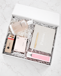 An open white gift box filled with crinkle paper and arranged beauty/self-care items, including a frosted bottle, a large gray notebook with the word 'Pause.' in white, a pink tube, a boxed item labeled 'Milk', and a small brown package, photographed from above on a light marble surface.