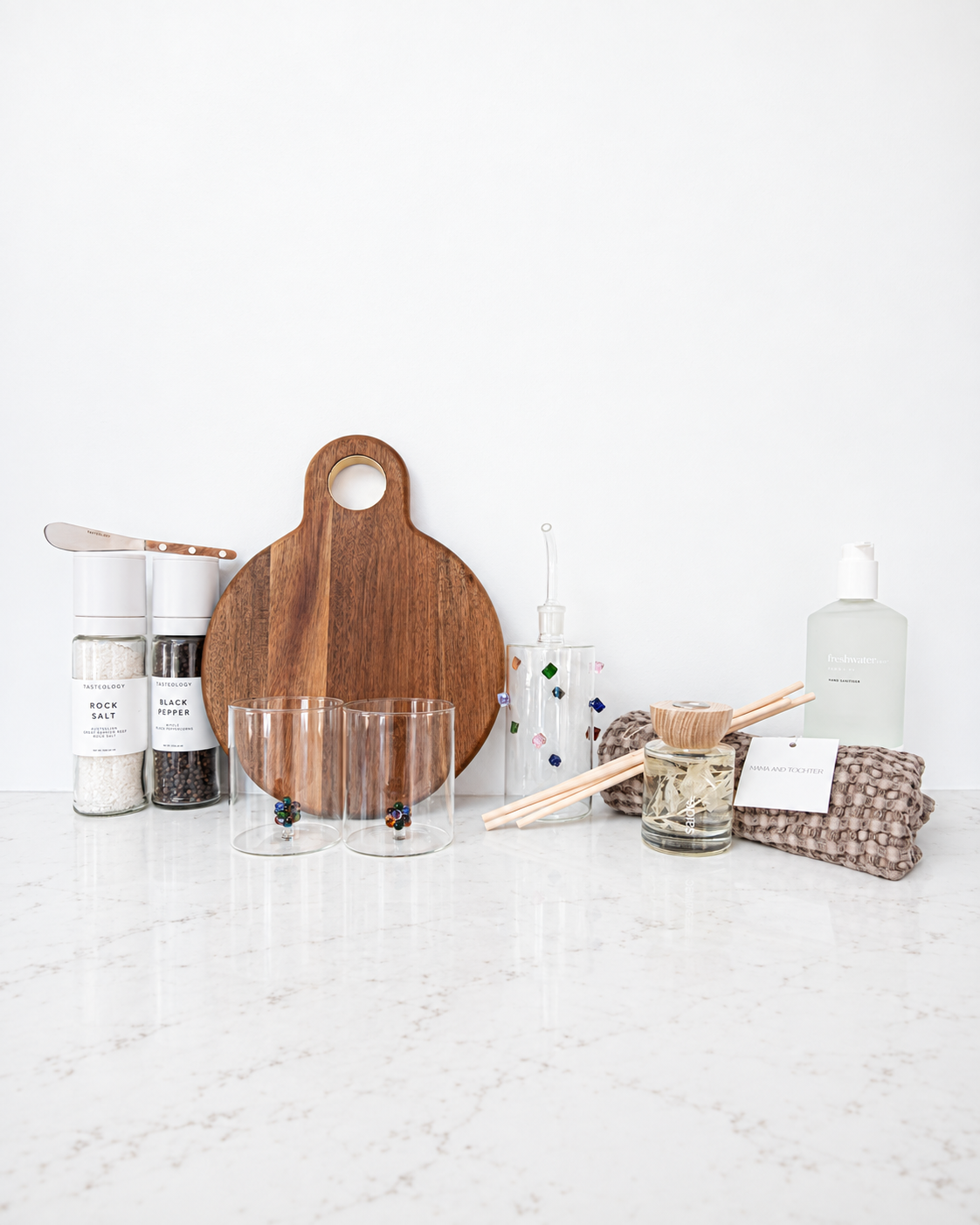A styled home goods and kitchen accessories set arranged on a white marble countertop against a white wall. The set includes a round wooden cutting board, two clear glass tumblers, two spice jars with white lids, a glass bottle with a dropper, a reed diffuser with wooden sticks, a patterned cloth, and a frosted bottle.