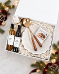 A holiday gift box set from featuring a white ceramic plate, gold-toned serving spoons with wooden handles, a small candle, and two bottles of olive oil and red balsamic vinegar, arranged on a marble surface with festive pine branches and lights.