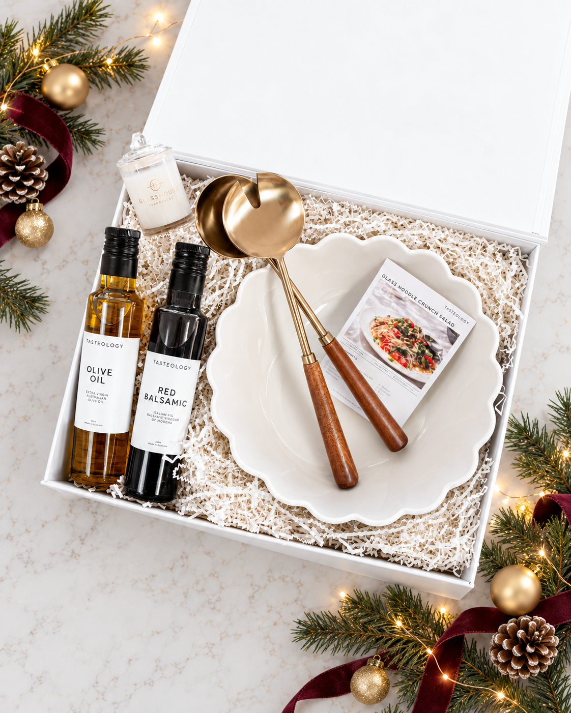 A holiday gift box set from featuring a white ceramic plate, gold-toned serving spoons with wooden handles, a small candle, and two bottles of olive oil and red balsamic vinegar, arranged on a marble surface with festive pine branches and lights.