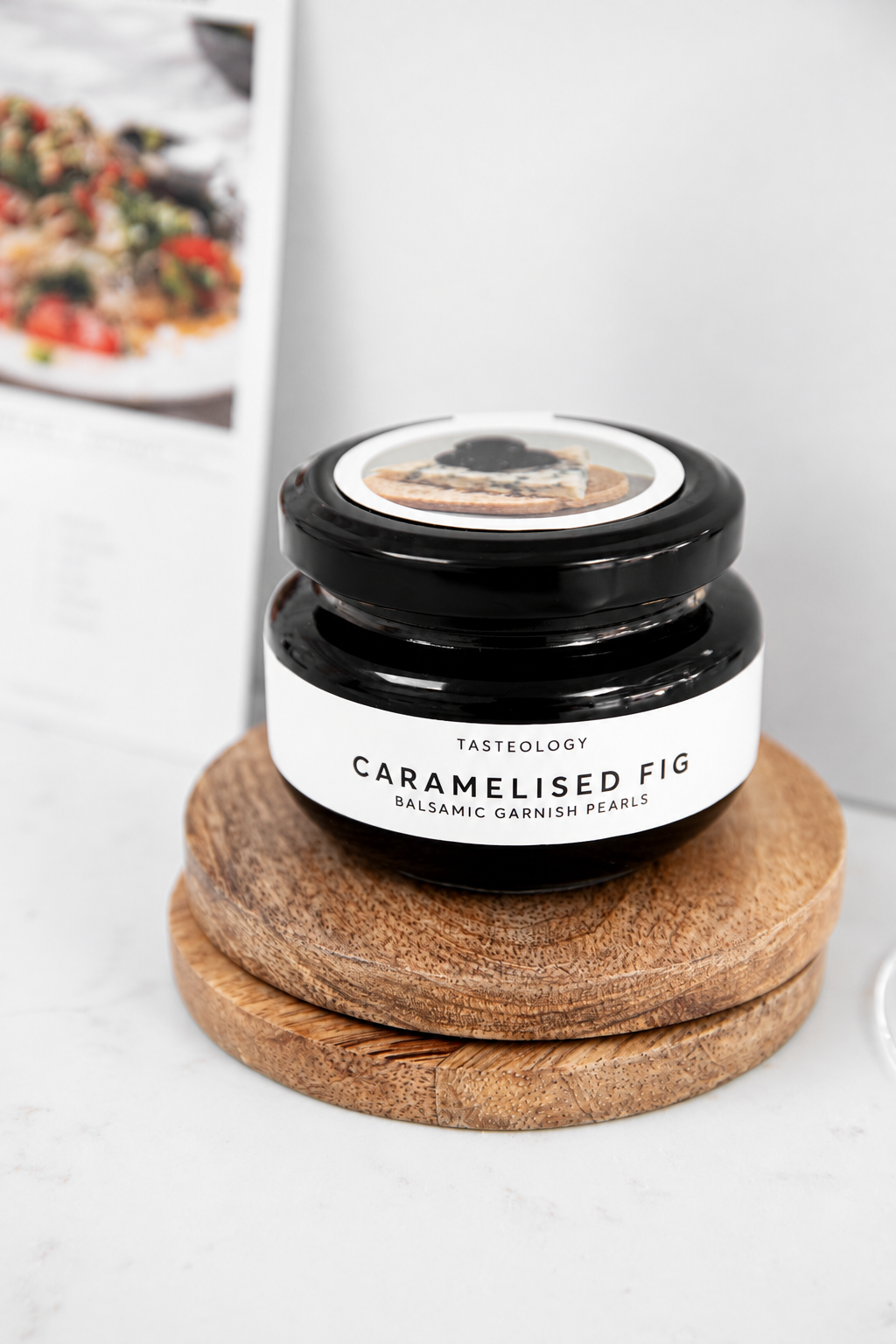 A small glass jar of Tasteology Caramelised Fig Balsamic Garnish Pearls sits on two stacked round wooden coasters against a light gray background.