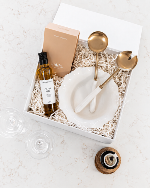 A curated gift box set on a white marble surface, featuring a bottle of Nautica olive oil, a beige 'saiide' box, a white ceramic bowl, and two gold-toned serving utensils with white handles nestled in shredded paper.