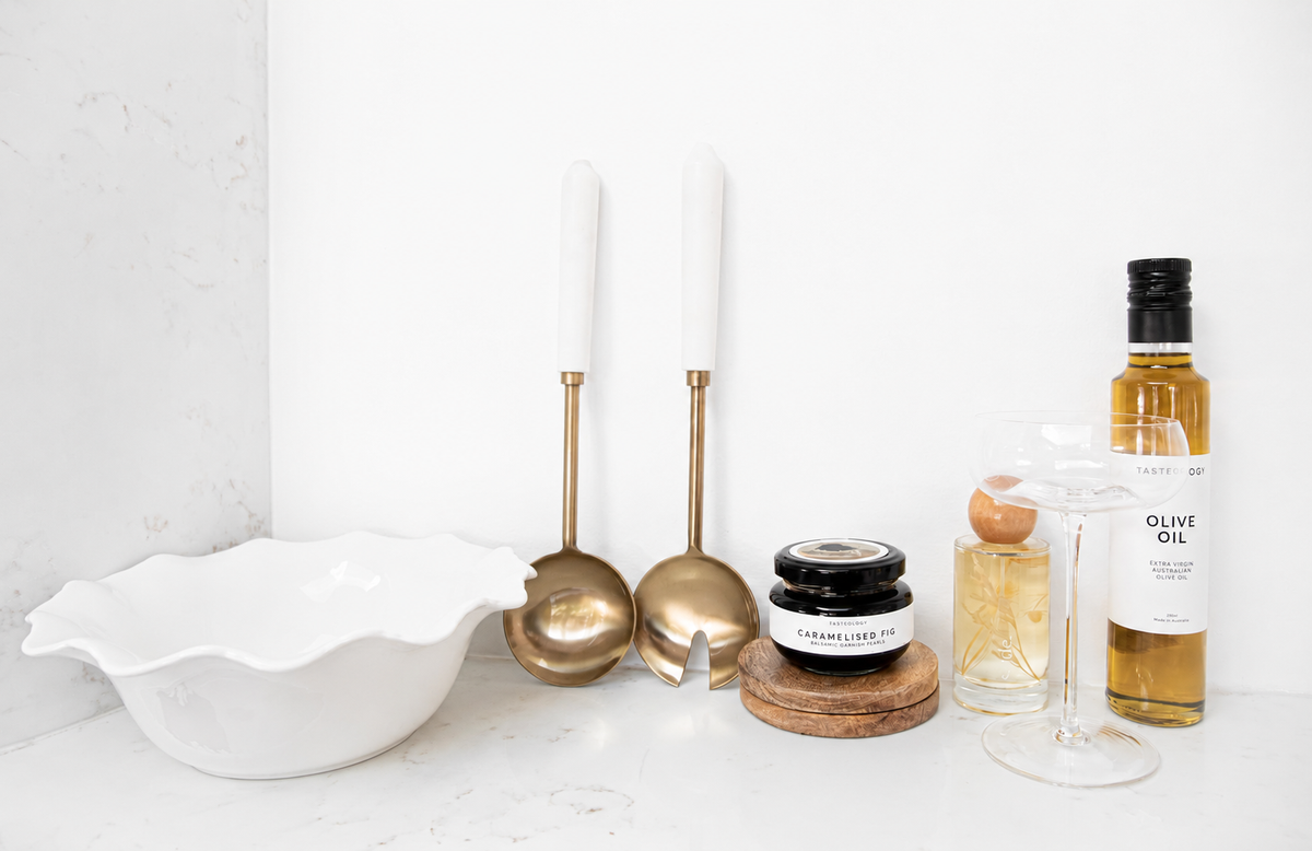 A styled kitchen countertop featuring a white scalloped ceramic bowl, two brass ladles with white handles, a small jar of Cranberry Sauce, a bottle of olive oil, and a wine glass against a white marble and plaster background.