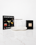 A bar-themed gift set on a white marble surface against a white wall, featuring a black cocktail book, two clear stemmed cocktail glasses on a white oval tray, and a black Arabian Nights box.