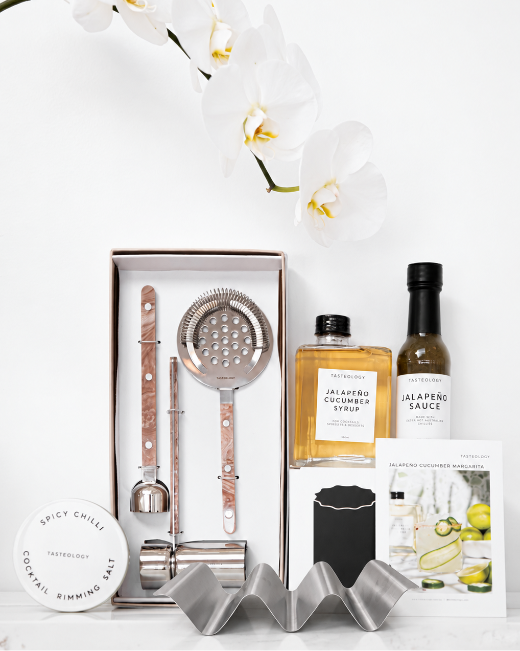 A curated cocktail and sauce gift set arranged on a white surface with white orchid flowers in the background. The set includes a boxed bar tool set, a bottle of jalapeño cucumber syrup, a bottle of jalapeño sauce, a round tin of spicy chili cocktail rimming salt, a small bottle, and printed cards, all styled cleanly against a white background.